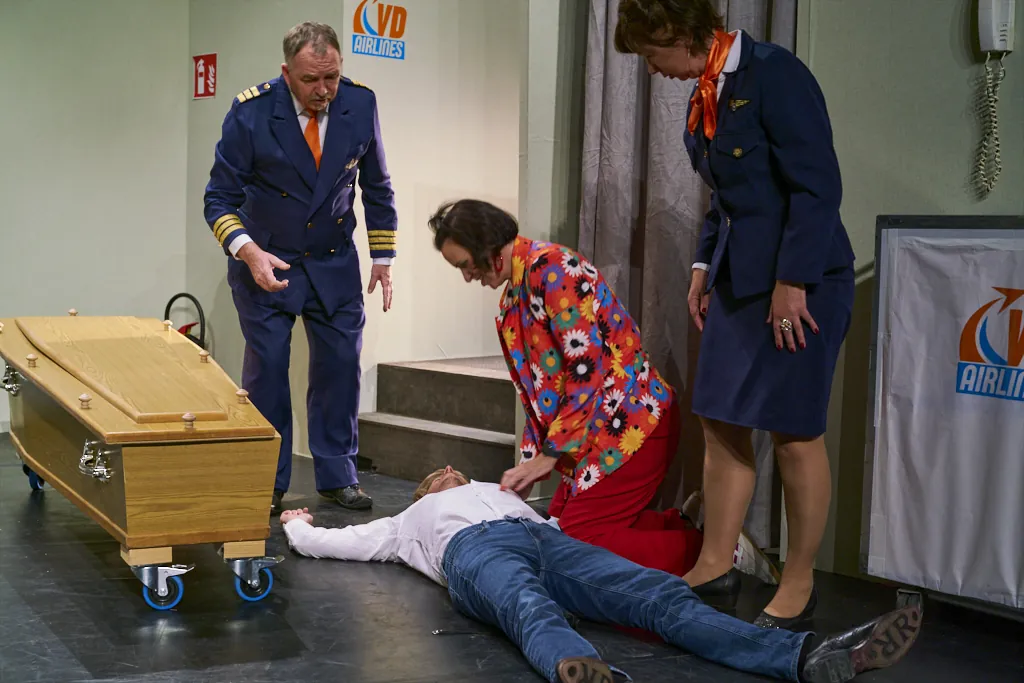 Evelyn buigt zich over een medespeler op de vloer, in een scène met de piloot en een stewardess